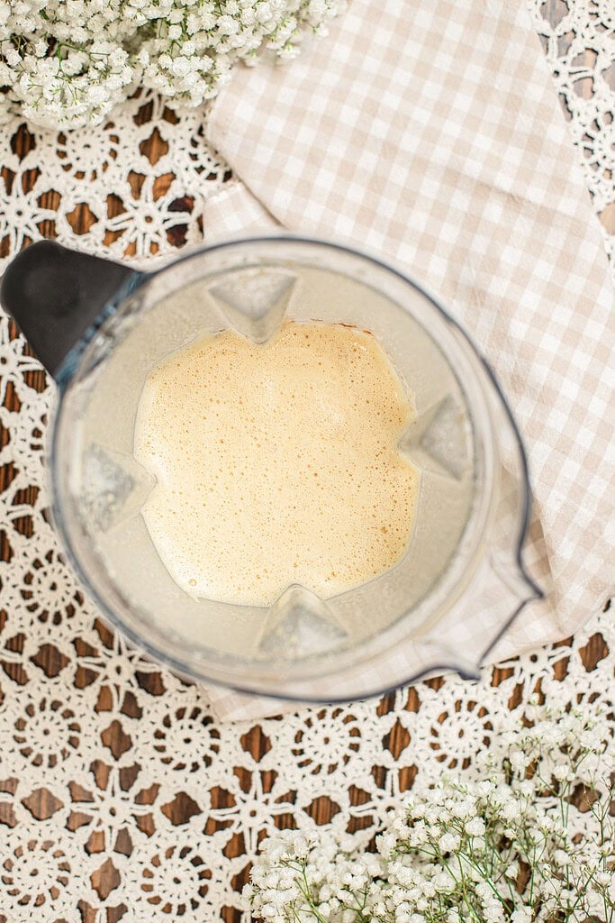 Overhead view of pale golden Dutch baby pancake batter blended smooth in a glass blender pitcher, resting on a beige checked cloth surrounded by baby's breath flowers.