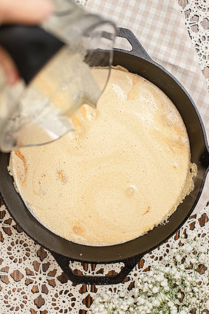 Blender pitcher pouring freshly milled flour Dutch baby batter into a hot cast iron skillet, with the batter spreading and foaming across the pan.