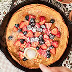 Overhead view of a baked Dutch baby pancake in a cast iron skillet being topped with fresh strawberries, blueberries, and blackberries, powdered sugar being sifted over the top, with a bowl of strawberries and raspberries nearby.