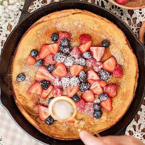 Overhead view of a baked Dutch baby pancake in a cast iron skillet being topped with fresh strawberries, blueberries, and blackberries, powdered sugar being sifted over the top, with a bowl of strawberries and raspberries nearby.