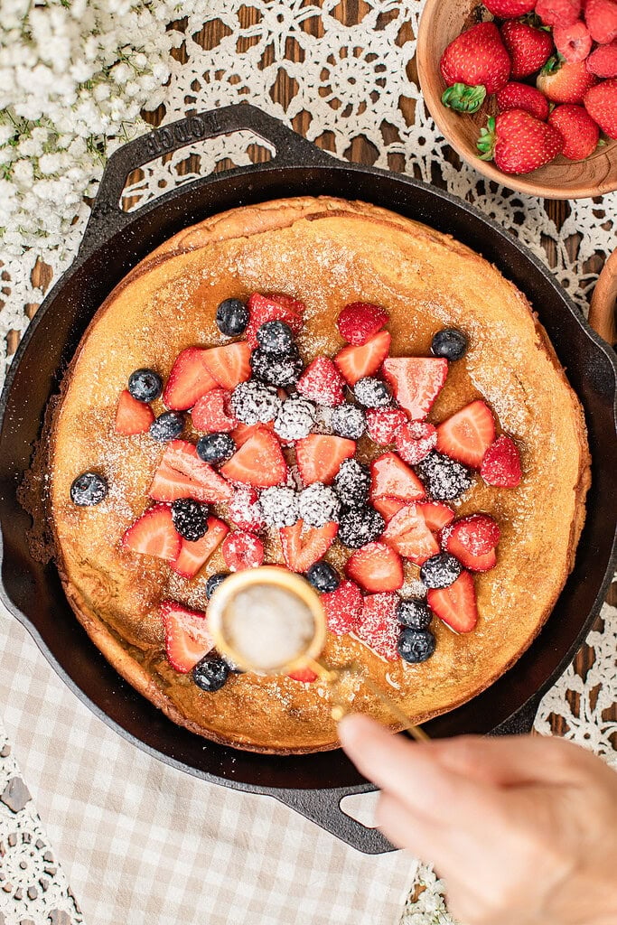 Overhead view of a baked Dutch baby pancake in a cast iron skillet being topped with fresh strawberries, blueberries, and blackberries, powdered sugar being sifted over the top, with a bowl of strawberries and raspberries nearby.