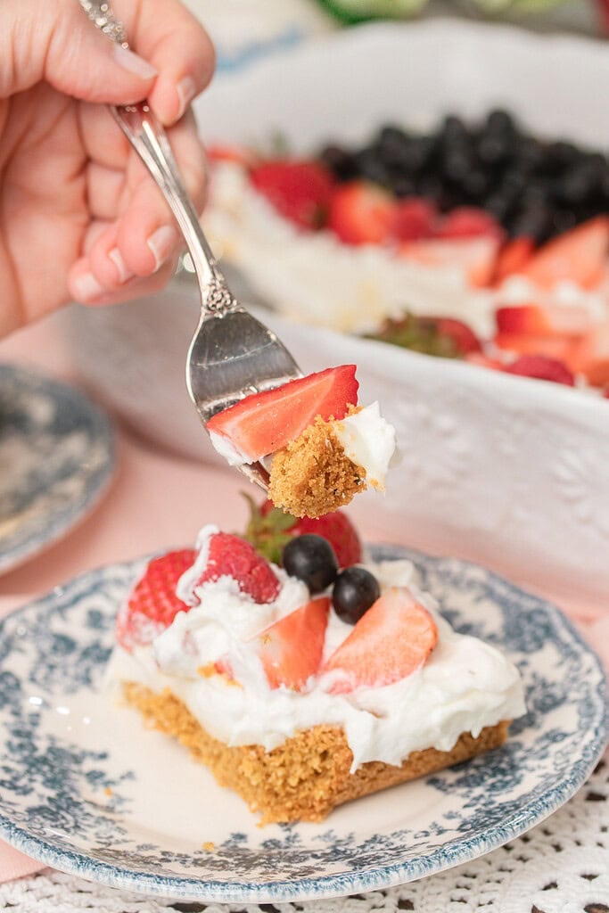 A hand holds a forkful of Fruit Pizza with Freshly Milled Flour showing a cookie crust, cream cheese frosting, and a strawberry slice, poised above a plated slice topped with strawberries and blueberries on a blue floral plate