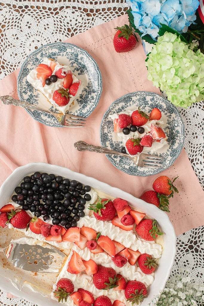 Two plated slices of Fruit Pizza with Freshly Milled Flour topped with whipped cream, strawberries, raspberries, and blueberries arranged in a flag pattern, resting beside the full sheet pan with a cake server, surrounded by blue and green hydrangeas