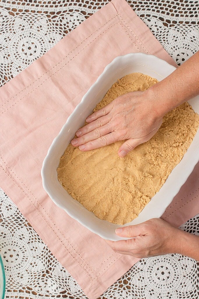 A hand pressing cookie dough into an even layer in a white rectangular baking dish to form the fruit pizza crust