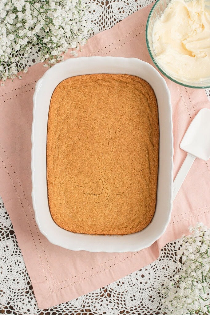 A baked golden cookie crust cooling in a white rectangular baking dish, with a bowl of whipped cream cheese frosting and baby's breath nearby