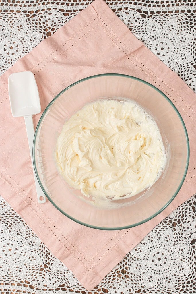 Whipped cream cheese frosting in a glass mixing bowl with a spatula resting beside it, ready to spread over the cooled cookie crust
