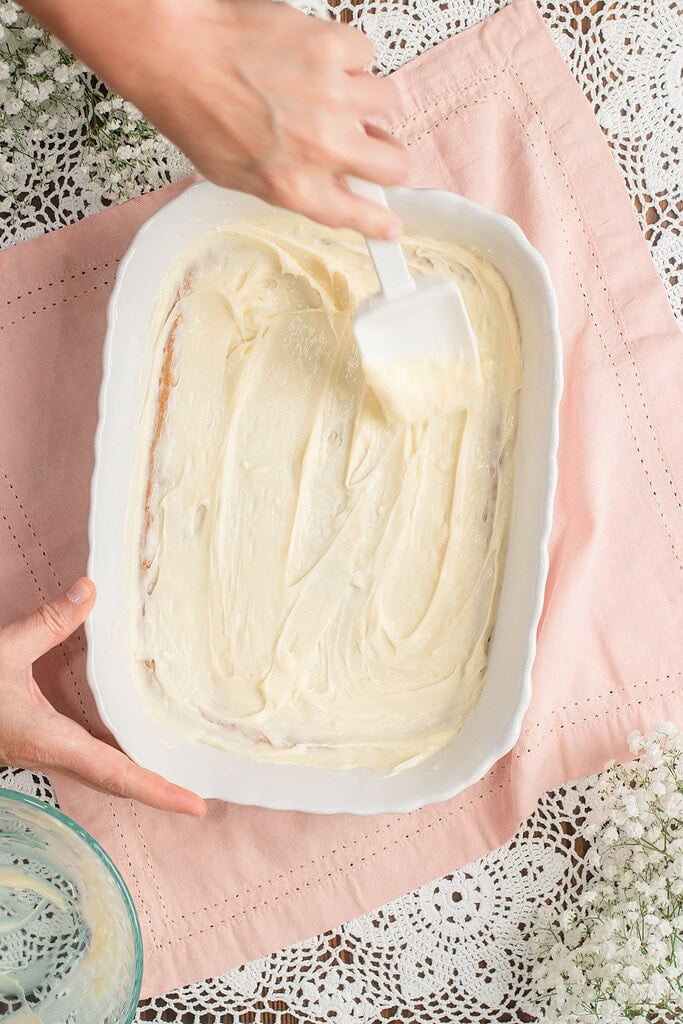 Hands spreading cream cheese frosting evenly over a baked cookie crust in a white rectangular baking dish