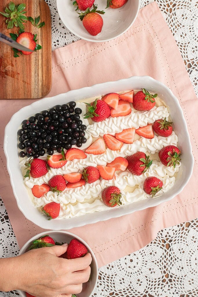 Overhead view of Fruit Pizza with Freshly Milled Flour being decorated in a flag pattern, with blueberries, sliced strawberries, and piped whipped cream stripes, a cutting board of hulled strawberries and a bowl of whole strawberries nearby