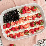 Finished Fruit Pizza with Freshly Milled Flour decorated as a flag design with blueberries, piped whipped cream, sliced strawberries, and raspberries, resting on pink linen with loose strawberries scattered around