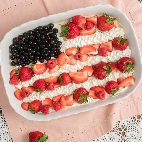 Finished Fruit Pizza with Freshly Milled Flour decorated as a flag design with blueberries, piped whipped cream, sliced strawberries, and raspberries, resting on pink linen with loose strawberries scattered around