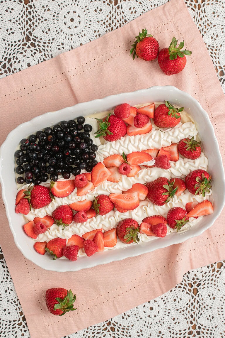 Finished Fruit Pizza with Freshly Milled Flour decorated as a flag design with blueberries, piped whipped cream, sliced strawberries, and raspberries, resting on pink linen with loose strawberries scattered around