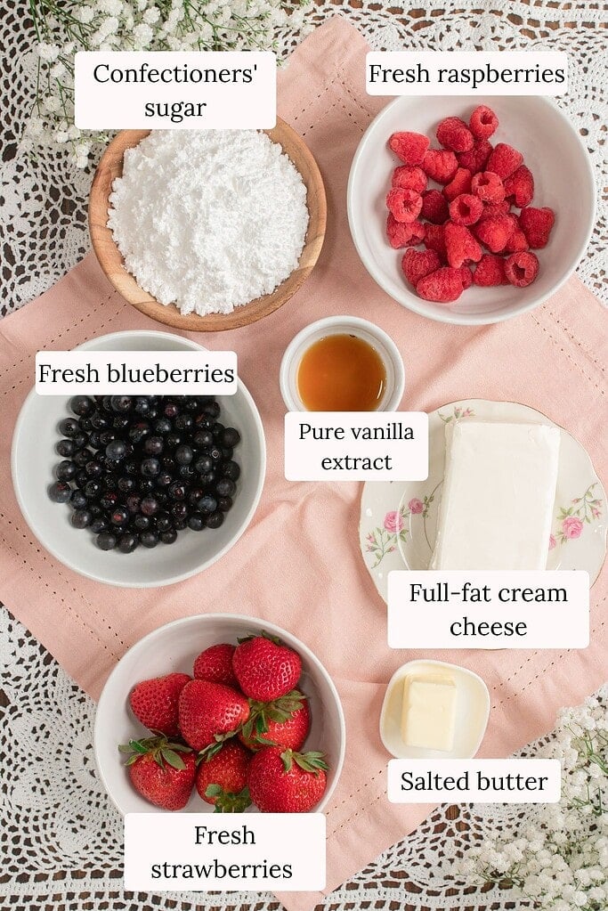 Overhead flat lay of fruit pizza toppings and frosting ingredients including confectioners sugar, fresh raspberries, fresh blueberries, pure vanilla extract, full-fat cream cheese, fresh strawberries, and salted butter arranged on pink linen with baby's breath