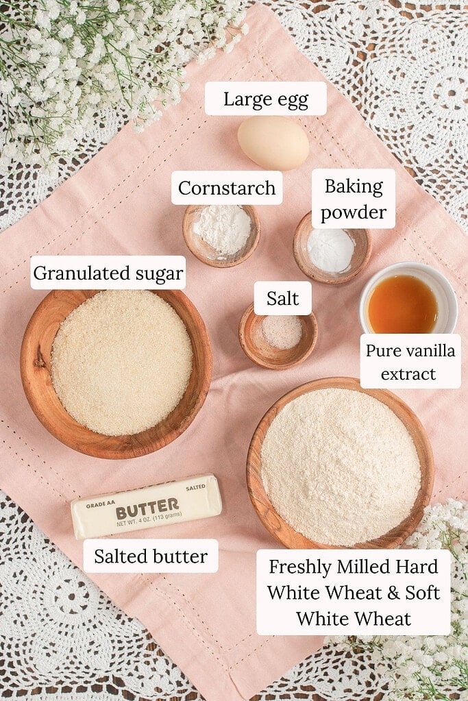 Overhead flat lay of cookie crust ingredients including a large egg, cornstarch, baking powder, granulated sugar, salt, pure vanilla extract, salted butter, and a bowl of freshly milled hard white and soft white wheat flour