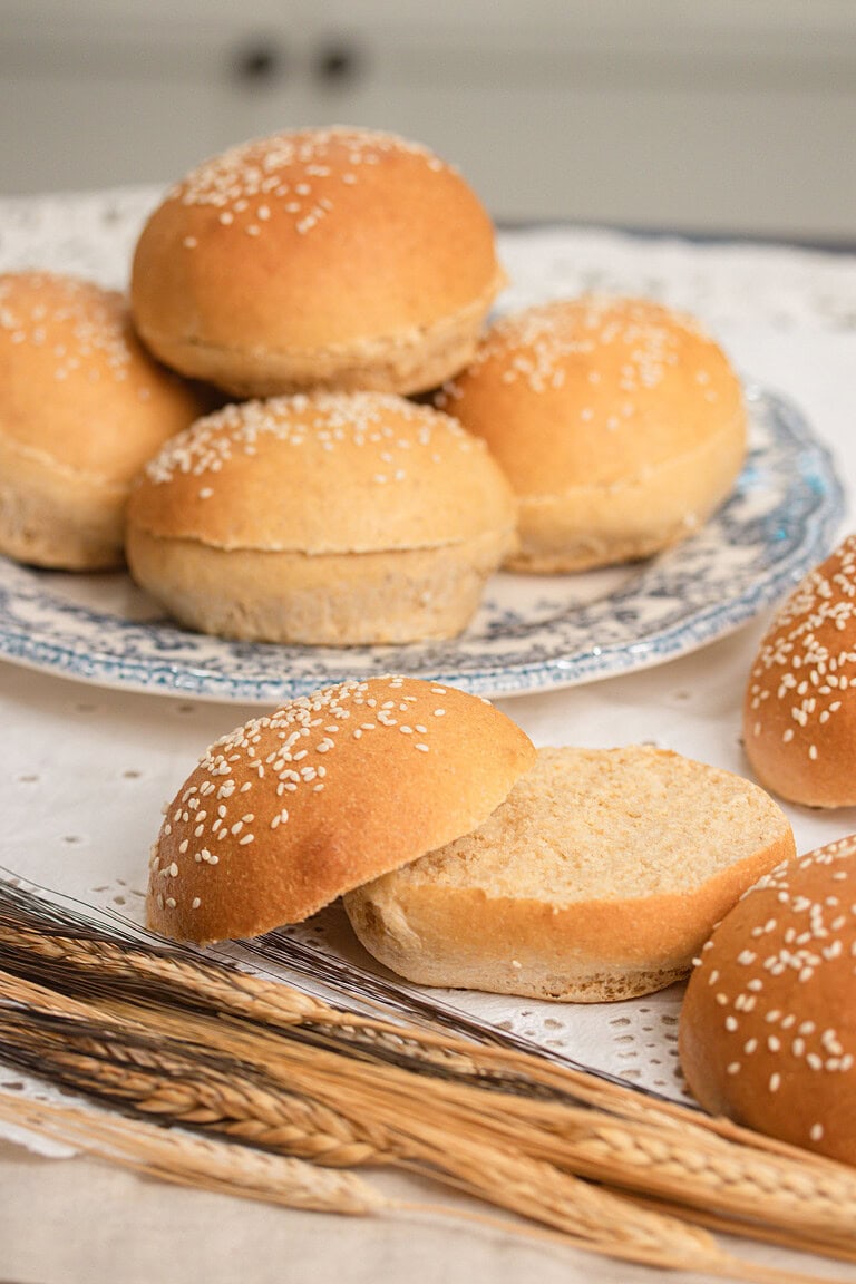 A stack of sesame seed topped Fresh Milled Flour Hamburger Buns on a blue floral plate, with one bun split open showing a soft interior, wheat stalks resting in the foreground