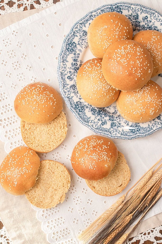 Several sesame seed topped Fresh Milled Flour Hamburger Buns arranged on a blue floral plate and scattered on linen, with two buns split open showing the soft crumb, wheat stalks in the foreground
