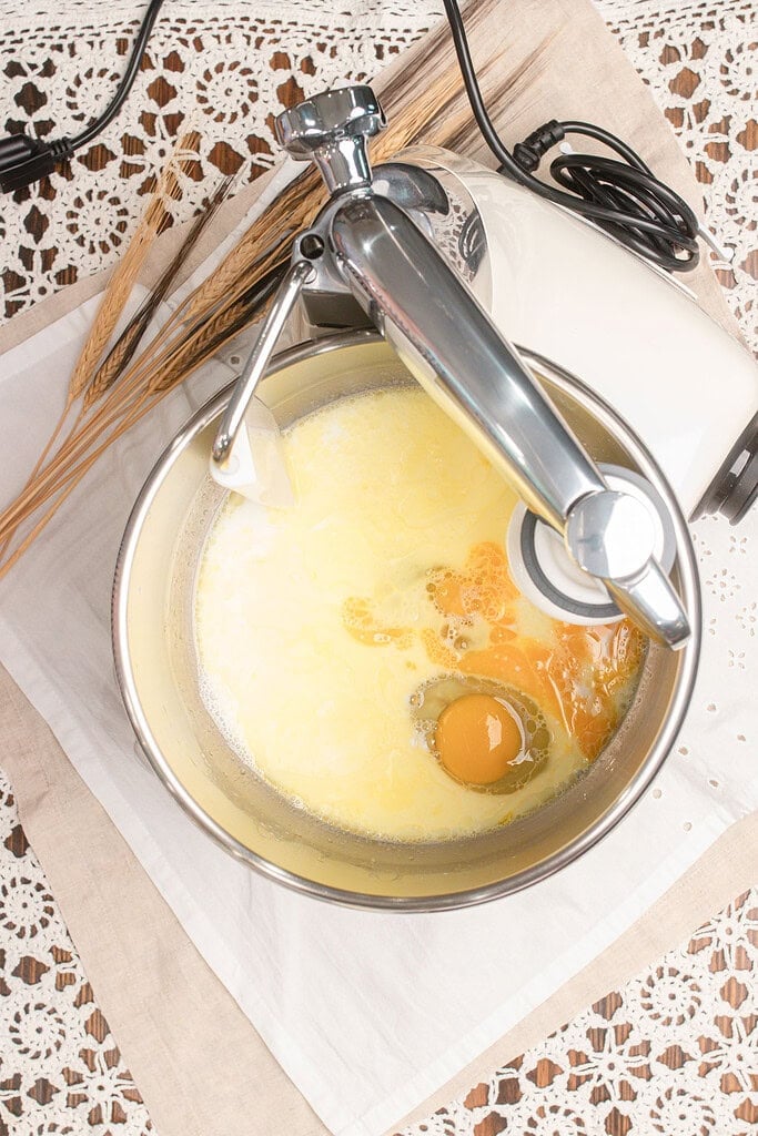 Overhead view of a stand mixer bowl with scalded milk, melted butter, and a cracked egg combined for the wet ingredients of hamburger bun dough