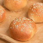 Close up of freshly baked sesame seed topped Fresh Milled Flour Hamburger Buns showing a golden crust on a parchment lined baking sheet
