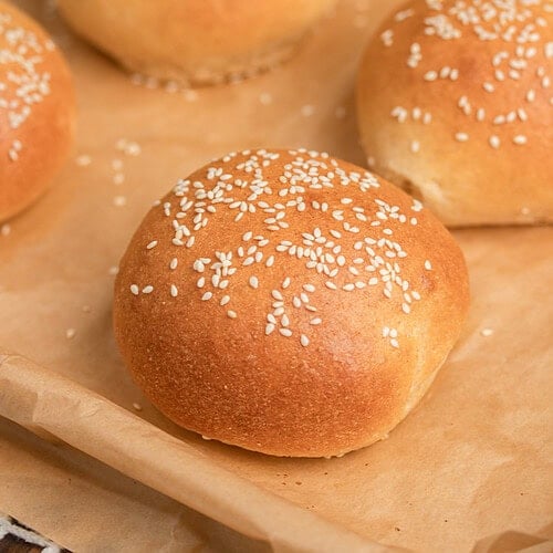 Close up of freshly baked sesame seed topped Fresh Milled Flour Hamburger Buns showing a golden crust on a parchment lined baking sheet