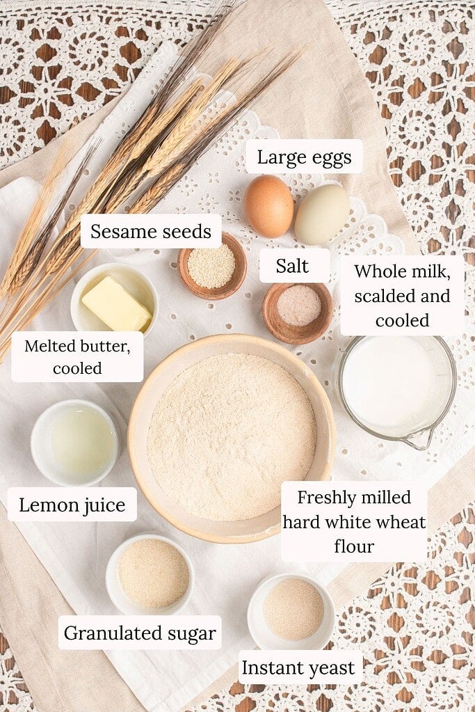 Overhead flat lay of hamburger bun ingredients including large eggs, sesame seeds, salt, whole milk, melted butter, lemon juice, granulated sugar, instant yeast, and a bowl of freshly milled hard white wheat flour arranged on linen with dried wheat stalks