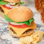 A fully loaded homemade cheeseburger with sesame seed bun, lettuce, tomato, onion, and cheddar cheese on a blue floral plate with waffle chips, another burger and iced tea glasses blurred in the background