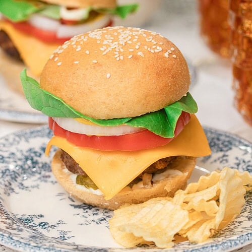 A fully loaded homemade cheeseburger with sesame seed bun, lettuce, tomato, onion, and cheddar cheese on a blue floral plate with waffle chips, another burger and iced tea glasses blurred in the background