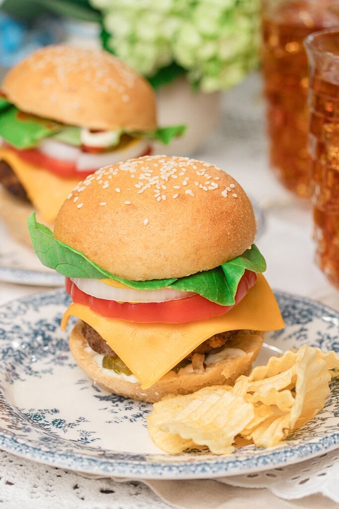 A fully loaded homemade cheeseburger with sesame seed bun, lettuce, tomato, onion, and cheddar cheese on a blue floral plate with waffle chips, another burger and iced tea glasses blurred in the background