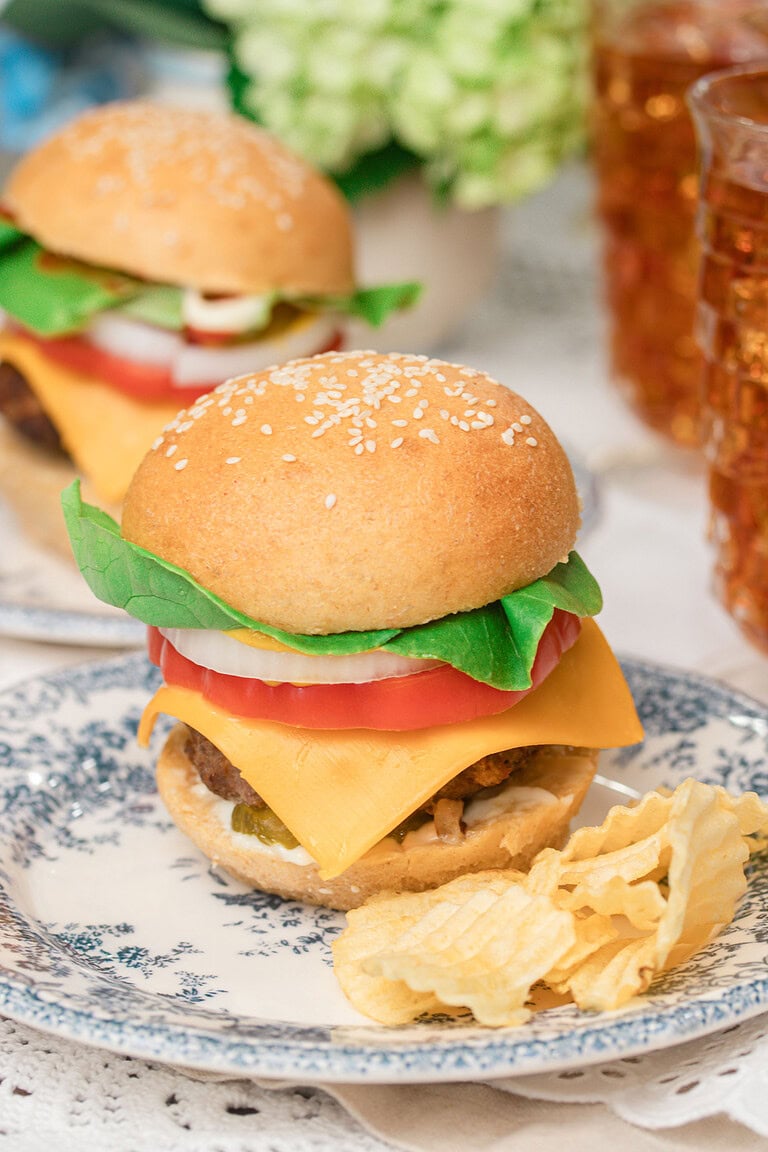 A fully loaded homemade cheeseburger with sesame seed bun, lettuce, tomato, onion, and cheddar cheese on a blue floral plate with waffle chips, another burger and iced tea glasses blurred in the background