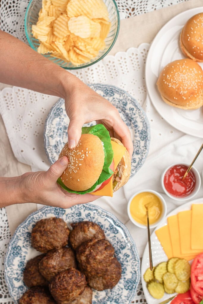 A hand holding a fully assembled cheeseburger with lettuce, tomato, and cheese, resting above a blue floral plate of cooked hamburger patties, with buns, condiments, cheese slices, pickles, and tomato slices arranged nearby
