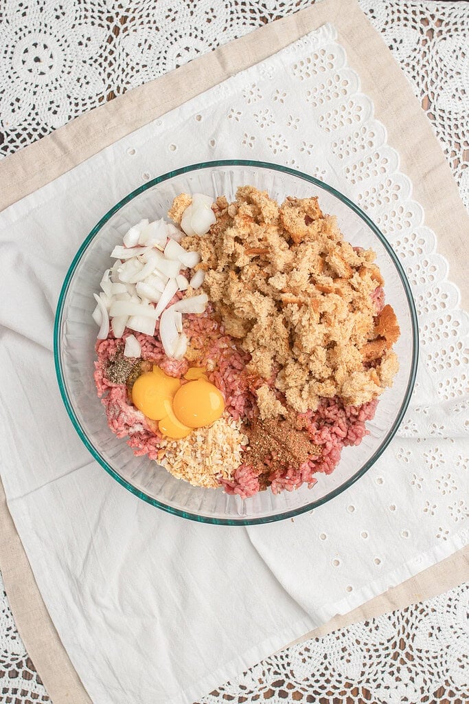 Overhead view of a glass mixing bowl with ground beef, diced onion, fresh breadcrumbs, egg yolks, toasted coconut flakes, and seasoning ready to be combined for homemade hamburger patties