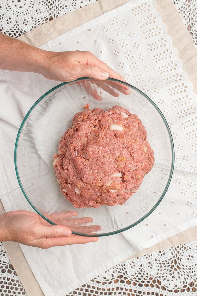 Hands holding a glass bowl filled with seasoned ground beef mixture formed into a mound, ready to be portioned into hamburger patties