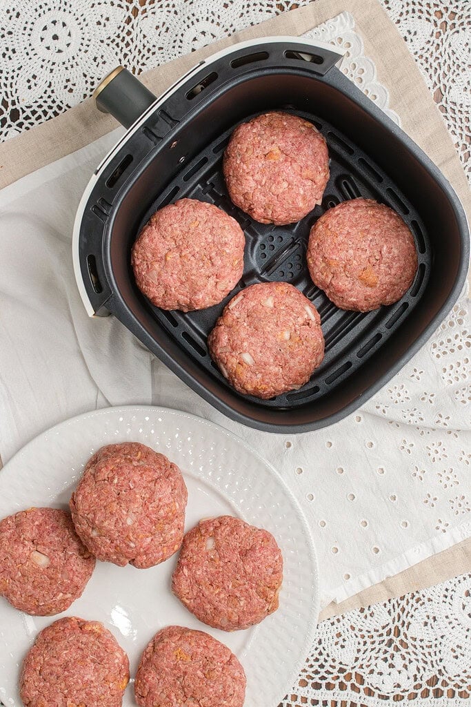Raw hamburger patties arranged in an air fryer basket alongside more shaped patties resting on a white plate nearby