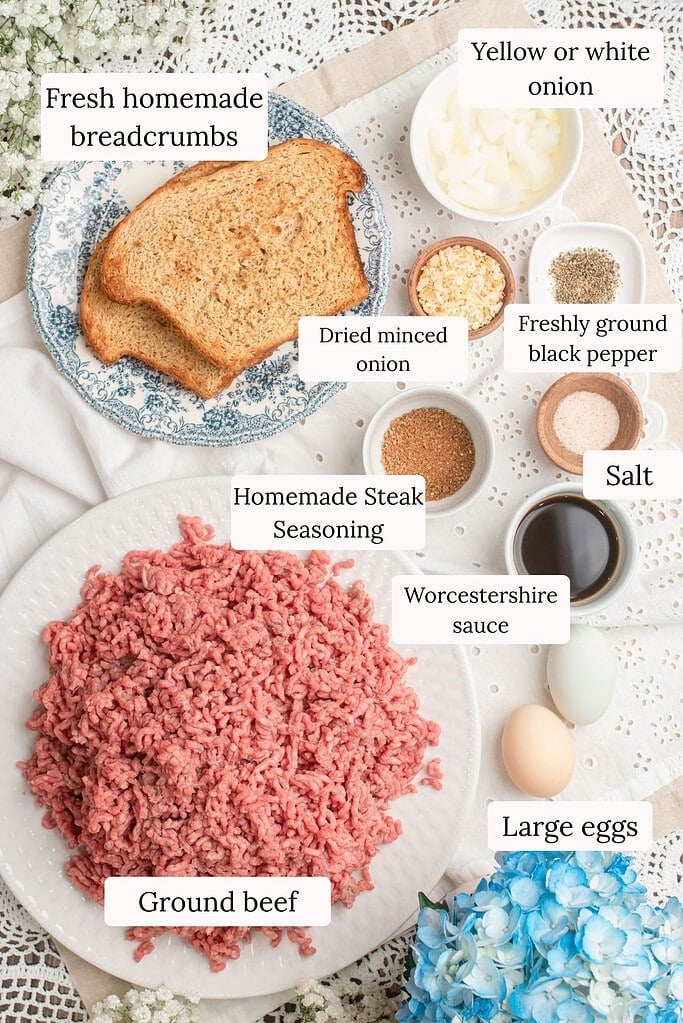 Overhead flat lay of hamburger patty ingredients including fresh homemade breadcrumbs, yellow onion, dried minced onion, freshly ground black pepper, homemade steak seasoning, salt, Worcestershire sauce, large eggs, and a plate of ground beef