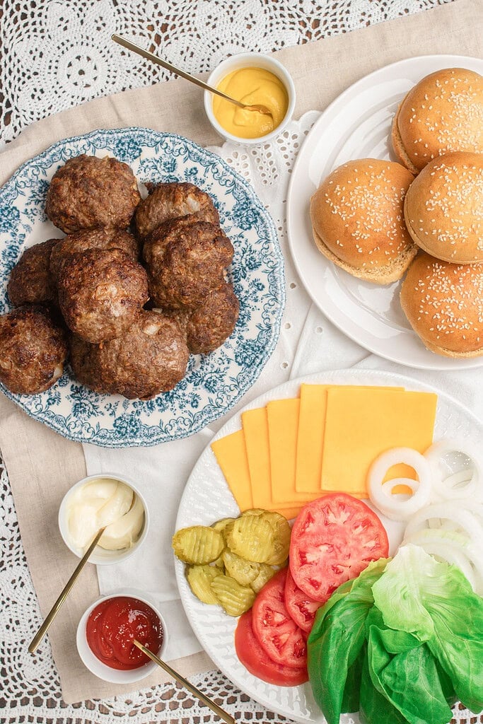 Cooked hamburger patties on a blue floral plate beside a plate of sesame seed buns, a plate of cheese slices, pickles, tomato, onion, and lettuce, with small dishes of ketchup, mustard, and mayonnaise