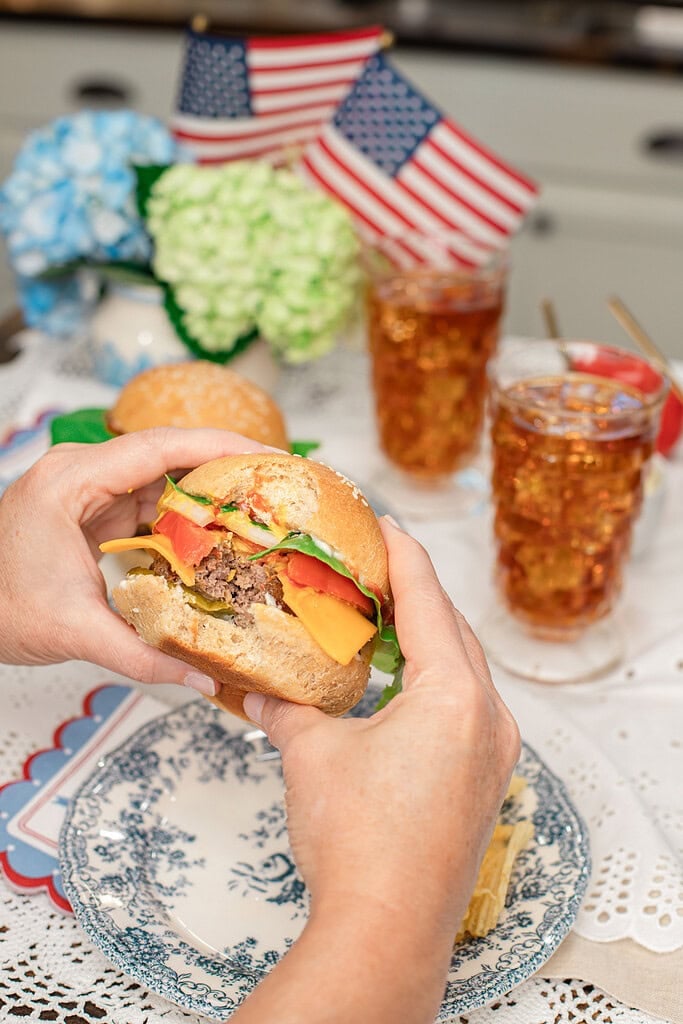 Close up of hands holding a loaded cheeseburger with melted cheese, tomato, lettuce, and onion, with iced tea glasses topped with American flag picks and hydrangeas blurred in the background