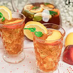 Homemade peach iced tea served over ice in vintage glasses, garnished with fresh peach slices, lemon slices, and mint, with a glass pitcher of iced tea in the background.