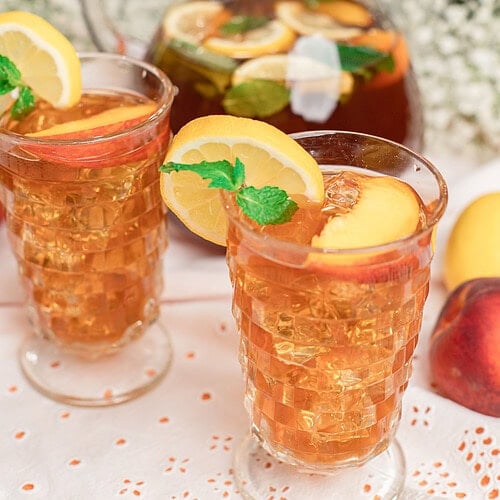 Homemade peach iced tea served over ice in vintage glasses, garnished with fresh peach slices, lemon slices, and mint, with a glass pitcher of iced tea in the background.