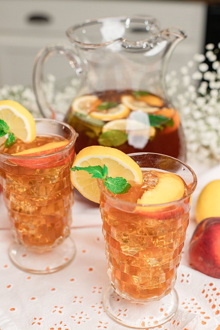 Homemade peach iced tea served over ice in vintage glasses, garnished with fresh peach slices, lemon slices, and mint, with a glass pitcher of iced tea in the background.