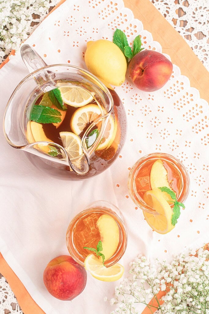Overhead view of homemade peach iced tea in a glass pitcher with fresh peaches, lemon slices, mint, and two glasses of iced tea served over ice on a white linen cloth.