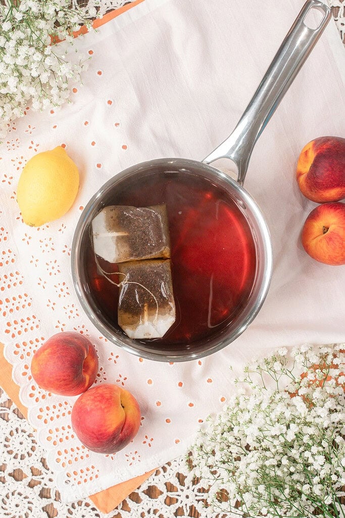 Black tea bags steeping in hot water in a saucepan during the first step of making homemade peach iced tea with fresh peaches and lemon nearby.