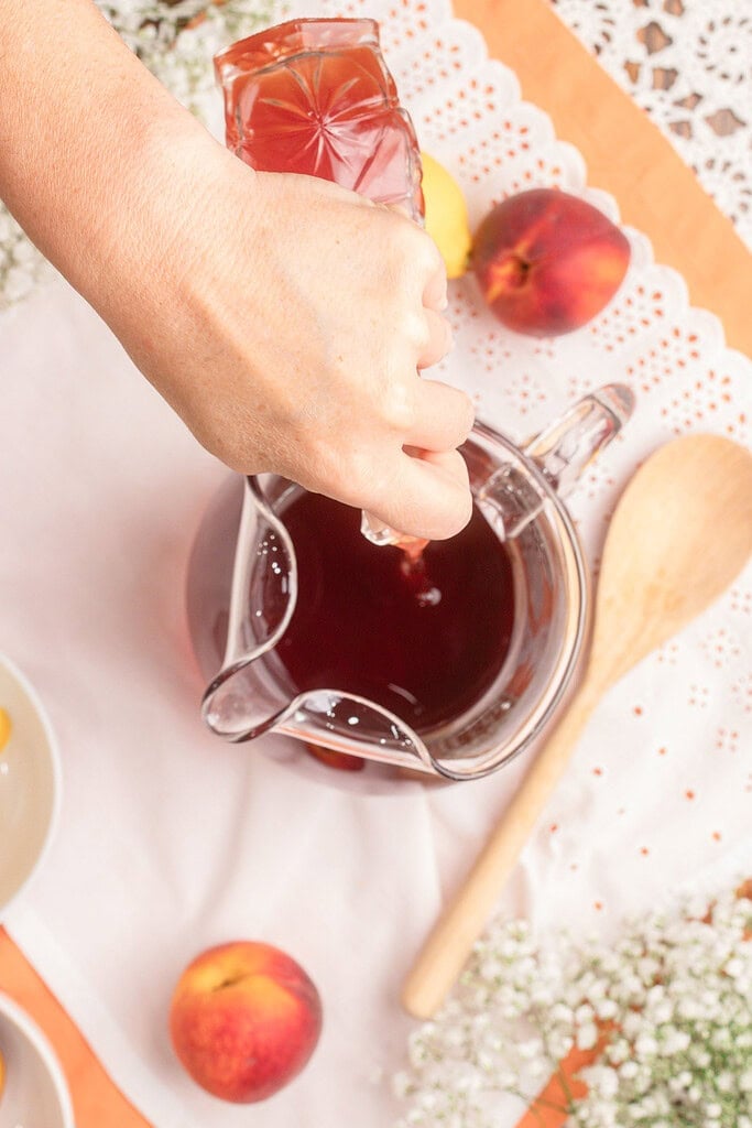 Pouring Peach Simple Syrup from a drinking glass into a glass pitcher while preparing homemade peach iced tea.