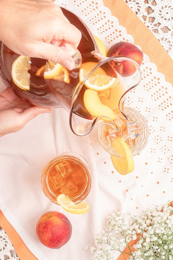 Pouring homemade peach iced tea from a glass pitcher into an ice-filled glass, with fresh peach slices, lemon slices, and mint floating in the pitcher.