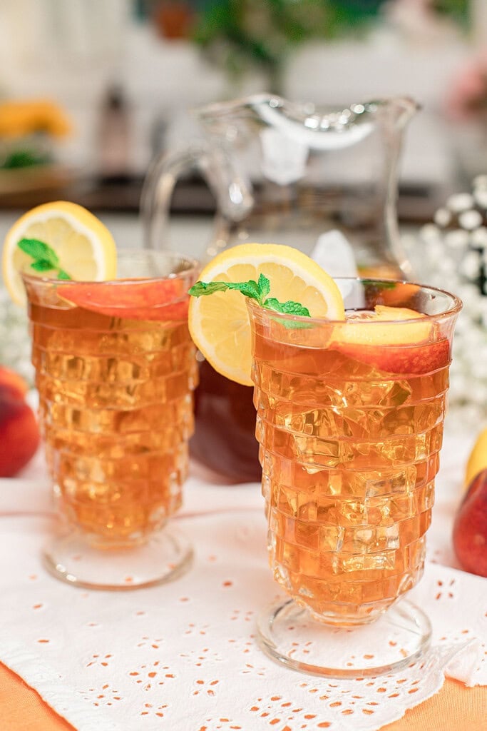 Two glasses of homemade peach iced tea served over ice with fresh peach slices, lemon wedges, and mint leaves, with a glass pitcher of iced tea in the background.