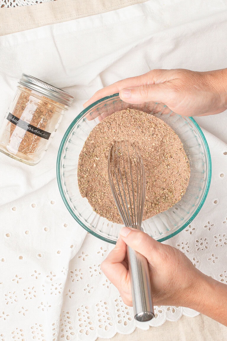 Homemade steak seasoning blend being whisked together in a glass mixing bowl with a labeled mason jar of the finished seasoning nearby