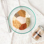 Unmixed spices for homemade steak seasoning arranged in sections in a glass bowl before whisking, with a whisk and mason jar nearby