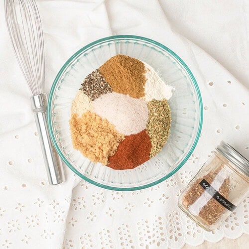 Unmixed spices for homemade steak seasoning arranged in sections in a glass bowl before whisking, with a whisk and mason jar nearby
