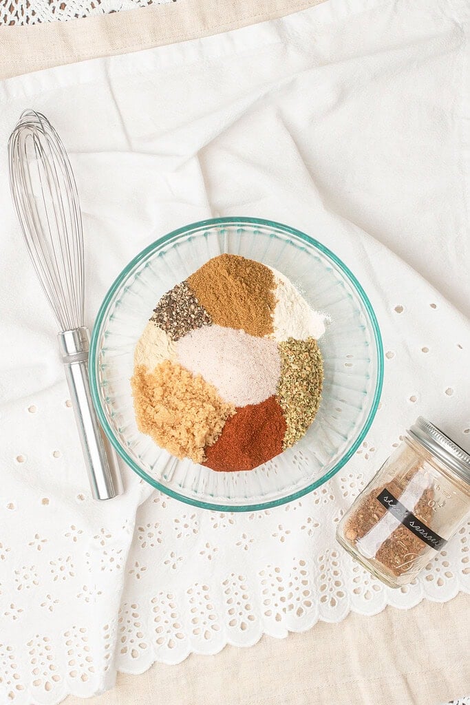 Unmixed spices for homemade steak seasoning arranged in sections in a glass bowl before whisking, with a whisk and mason jar nearby