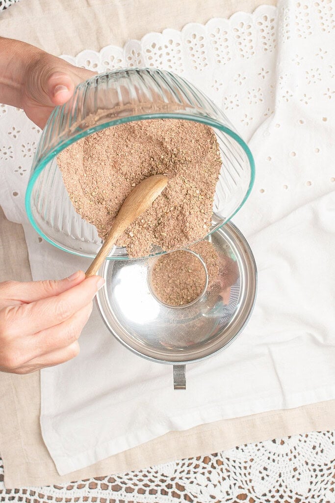 Homemade steak seasoning being poured from a glass bowl into a metal bowl with a wooden spoon guiding the transfer