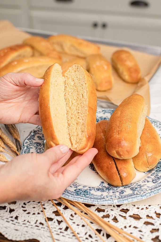 A hand holding open a sliced hot dog bun made with freshly milled flour showing its soft interior, with more baked buns and a plated stack in the background