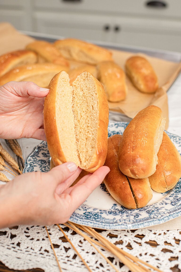 A hand holding open a sliced hot dog bun made with freshly milled flour showing its soft interior, with more baked buns and a plated stack in the background
