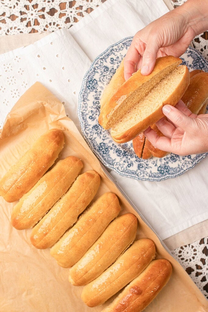 A hand holding a split hot dog bun made with freshly milled flour above a tray of baked buns and a plate stacked with more buns